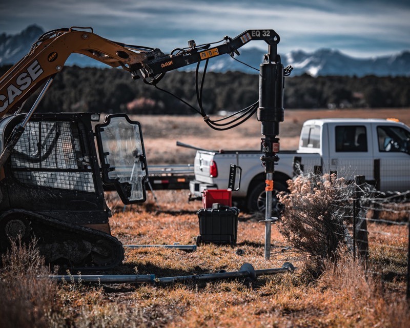 Compact track loader driving a helical pier on a rural lot, Site Safe work truck behind