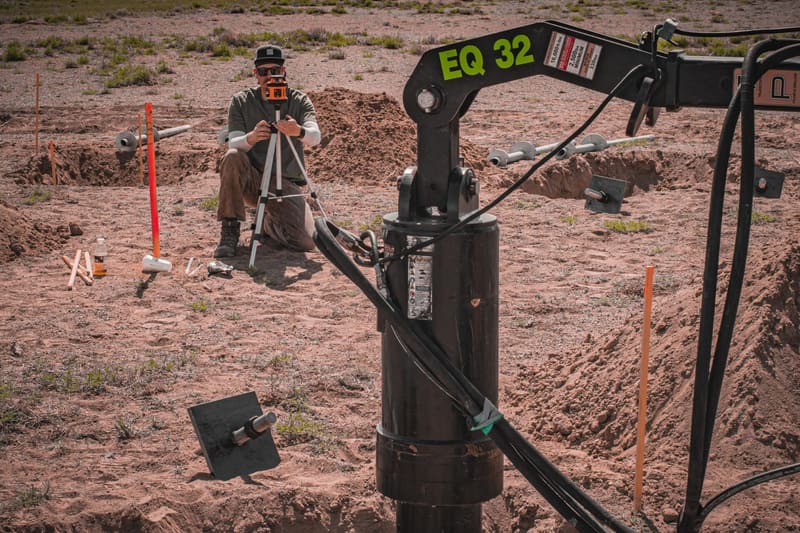 Site Safe installer with a pier drive head and survey stakes on an open site