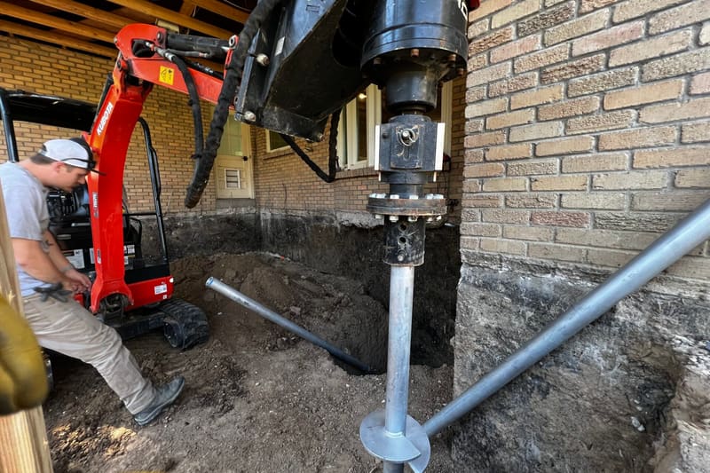 Helical pier being driven beside the brick foundation of a house