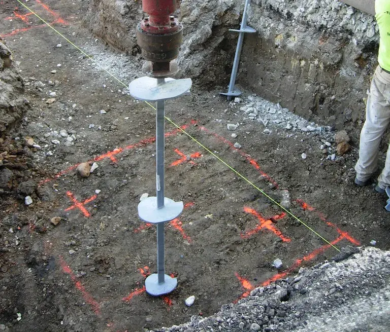 Helical pile with two helix plates being screwed in by a hydraulic drive head inside an excavation marked with paint layout lines