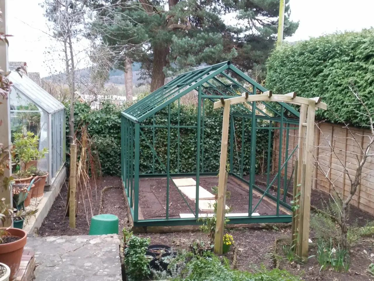 Green aluminum greenhouse frame under construction on a timber sleeper base in a garden, before the glazing is fitted