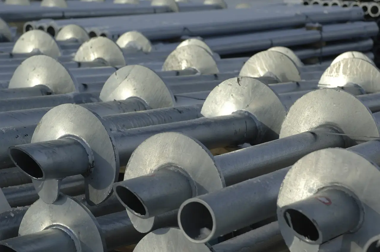Galvanized helical piles with single helix plates stacked on a job site, part of a helical pile specification's product requirements