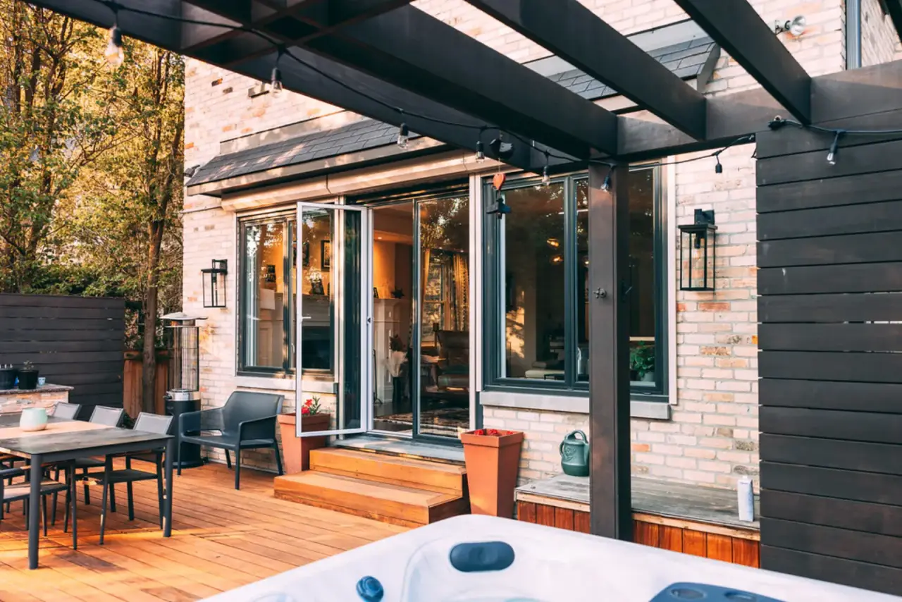 Hot tub on a hardwood deck beside a brick house, with a dark timber pergola overhead and a dining table nearby