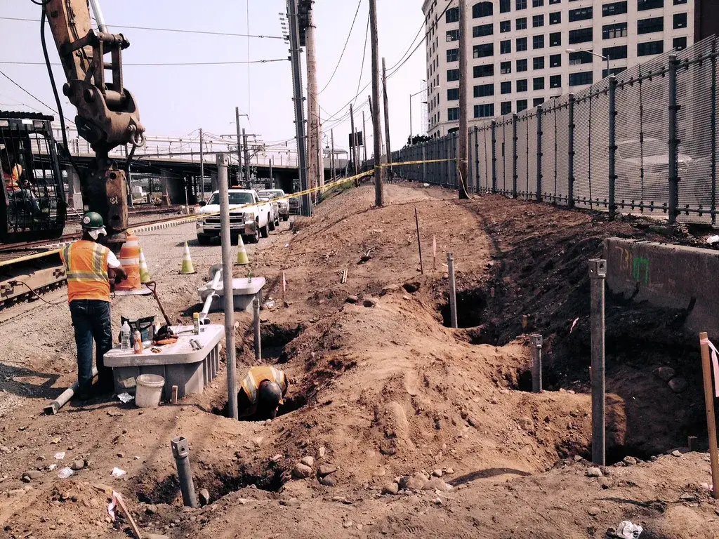A row of steel helical pile shafts installed in a trench for a new equipment foundation, with a crew working beside them