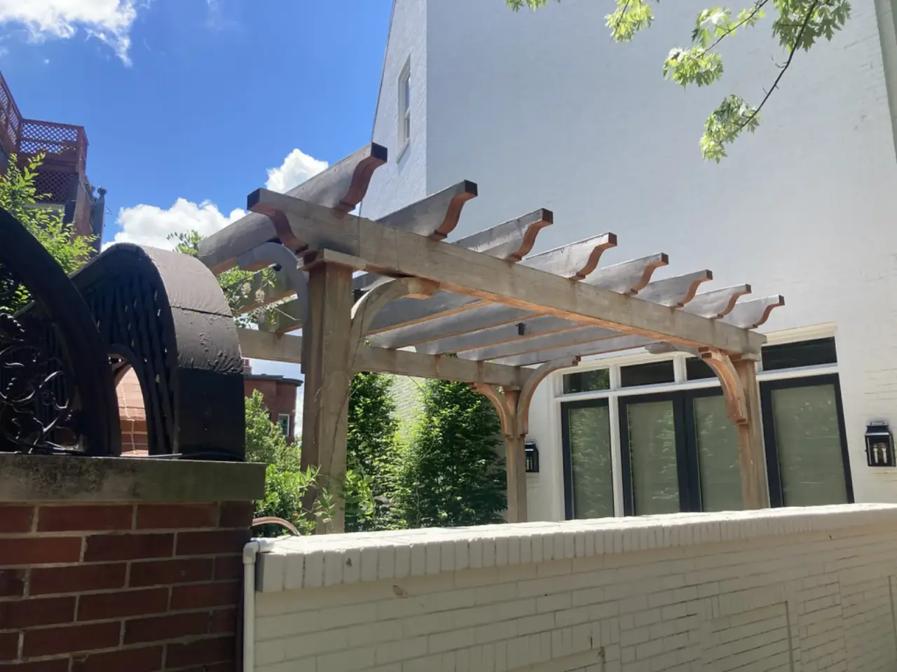 Timber pergola with a thick beam and curved-end rafters standing at the corner of a white brick house
