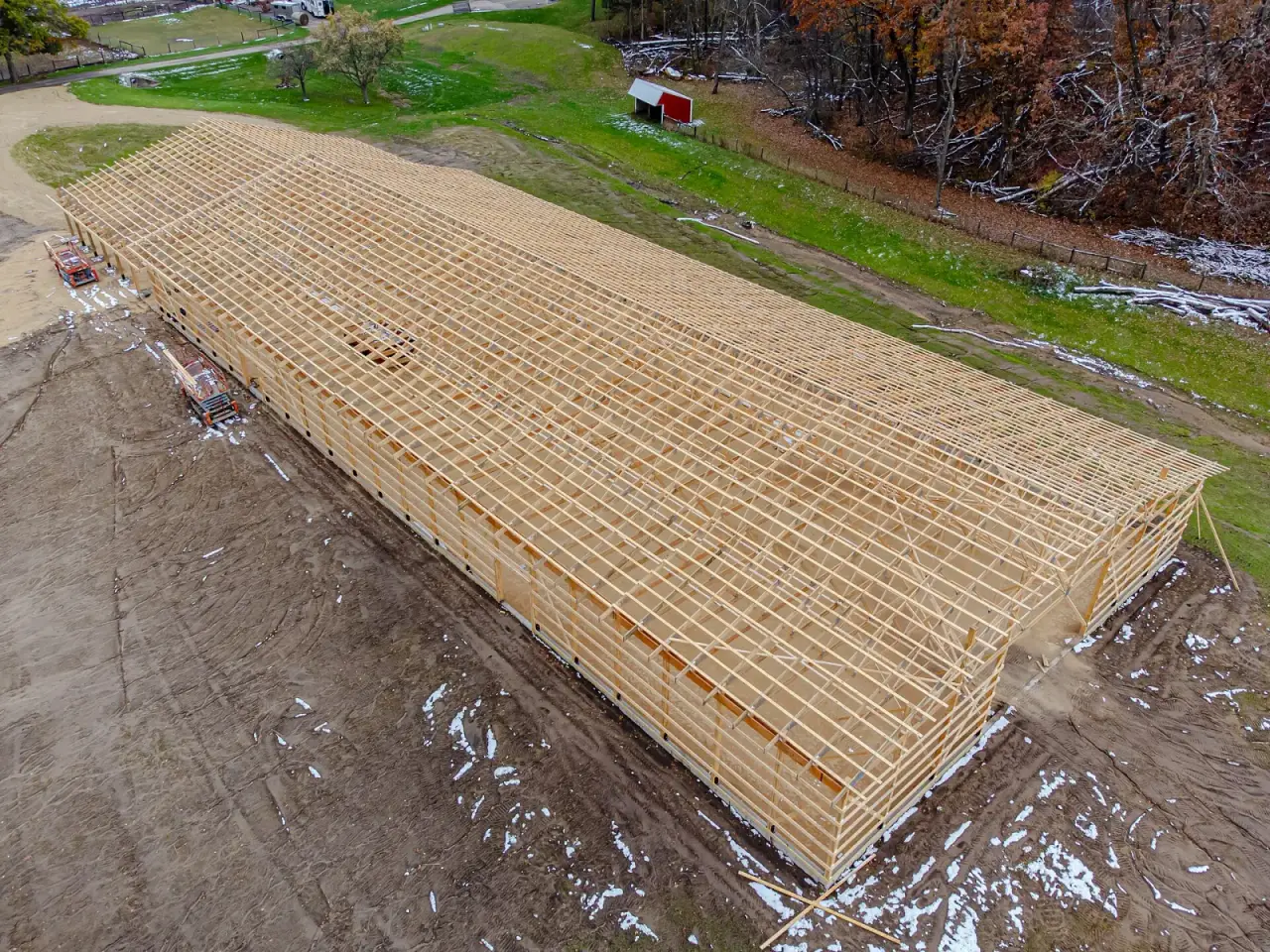 Aerial view of a long post-frame building with its posts, girts and roof trusses framed on a bare dirt site