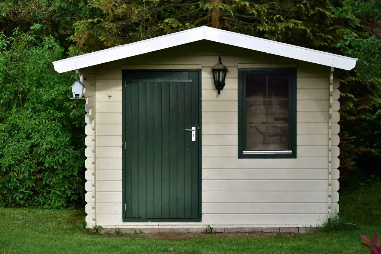 Small wooden garden shed with a green door and window sitting on a low base in a grassy yard